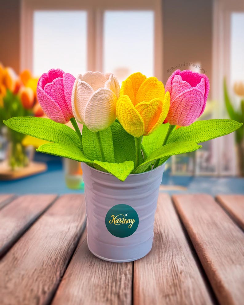 May include: A handmade crochet flower arrangement featuring four tulips in a white metal pot. The tulips are pink, cream, and yellow, with green leaves. The pot has a circular sticker with the word "Kasınkay".