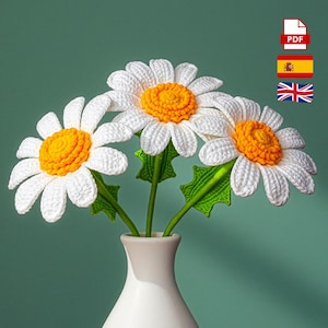 Puede incluir: Un ramo de tres margaritas de ganchillo en un jarrón blanco. Las margaritas tienen pétalos blancos y centros de color naranja brillante. Los tallos y las hojas verdes sostienen las flores. El jarrón es blanco y el fondo es de color verde azulado sólido.