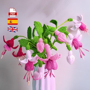 Puede incluir: Un ramo de flores de fucsia de ganchillo en un jarrón rosa pálido. Las flores son en tonos de rosa y blanco, con tallos y hojas verdes. La imagen incluye un icono PDF, la bandera española y la bandera británica.