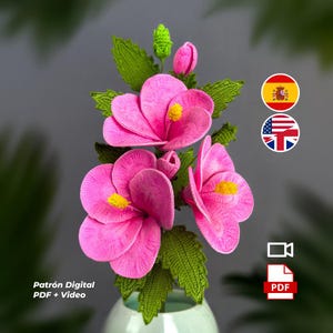 Puede incluir: Un ramo artesanal de flores rosas de ganchillo con centros amarillos y hojas verdes. Las flores están dispuestas en un jarrón verde claro. La imagen incluye el texto "Patrón Digital PDF + Video".