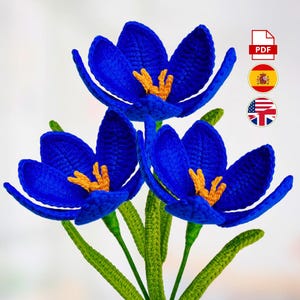 Puede incluir: Tres flores de ganchillo hechas a mano con pétalos de color azul vibrante y centros amarillos. Los tallos y las hojas son de color verde brillante. Las flores están dispuestas en un ramo. La imagen incluye un icono PDF y banderas de España, Estados Unidos y Reino Unido.