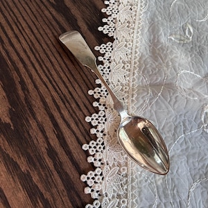 May include: A silver spoon with a long handle and a rounded bowl, resting on a dark wooden surface. The spoon is next to a white lace trim and a sheer, embroidered fabric.