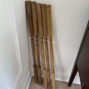 May include: A set of five vertical wooden balusters featuring a natural light brown finish. Each baluster has a square top and bottom section with a central turned spindle design, showcasing visible wood grain and a smooth, matte texture throughout.
