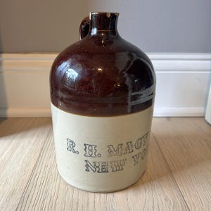 May include: A vintage brown and beige stoneware jug with a handle. The top portion is a glossy brown, while the bottom is beige with the text "R. H. Macy New York" in black lettering. The jug has a rounded body and a narrow neck.