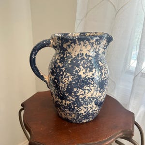 May include: A ceramic pitcher featuring a dark blue and white speckled sponge-painted pattern. The pitcher has a rounded body, a curved handle on the side, and a small pouring spout at the top rim, showcasing a glossy finish throughout the exterior.