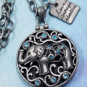 May include: A silver necklace with a round pendant featuring an elephant design with blue rhinestones. The pendant is attached to a silver chain. A small silver tag hangs from the chain with the text "love courage adventure" inscribed on it.