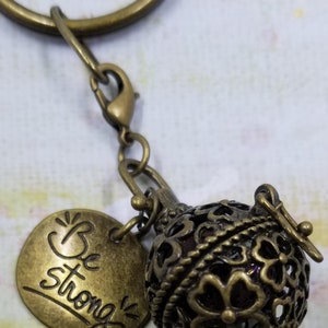 Puede incluir: Llavero de color bronce antiguo con un dije redondo de estilo filigrana y un dije de disco más pequeño. El dije de disco tiene las palabras "Be strong" en una fuente manuscrita. El dije de filigrana es una esfera con recortes florales.