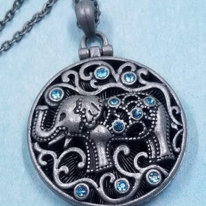 Puede incluir: Un collar de plata con un colgante redondo que presenta un diseño de elefante con diamantes de imitación azules. El colgante tiene un diseño de filigrana alrededor del elefante.