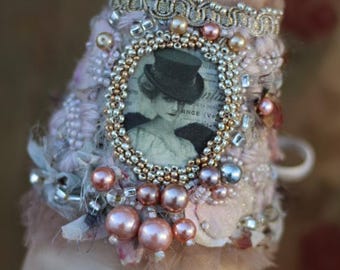 May include: A handmade cuff bracelet with a vintage aesthetic. The bracelet features a central oval portrait, surrounded by gold beads. Pink and silver beads and pearls adorn the cuff, which is made of textured fabric.