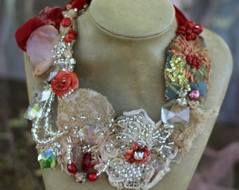 Puede incluir: Un collar artesanal de estética vintage. Cuenta con una cinta de terciopelo rojo, adornada con encaje, cuentas y flores de tela en tonos rojos, beige y plateados. El punto focal es un gran diseño floral ornamentado.