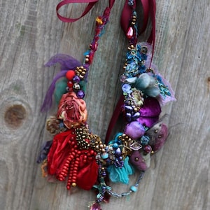 May include: A colourful, handmade necklace featuring a variety of beads, fabric and embellishments. The necklace includes a red velvet ribbon, a silk rose and a range of beads in shades of blue, purple and gold.