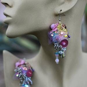 Può includere: Orecchini fatti a mano, colorati, con un design artistico unico. Gli orecchini presentano un mix di tessuti, perline e paillettes, nei toni del rosa, viola, giallo e blu. Pendono da ganci color argento, creando un accessorio stravagante e accattivante.