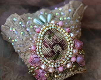 May include: A beige lace cuff bracelet adorned with iridescent rhinestones, pearls, and pink roses. The centerpiece features an oval embroidered design framed by gold-toned beads and crystals. A delicate, vintage-inspired accessory.