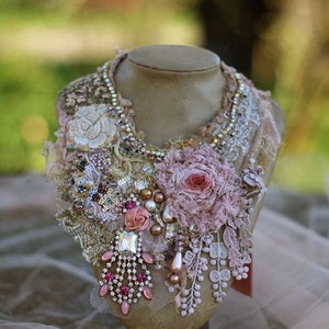 Può includere: Una collana delicata e stratificata con fiori di tessuto rosa e bianco, perle, strass e pizzo. La collana è esposta su un busto di manichino.