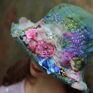 Könnte beinhalten: Ein grüner Schlapphut, verziert mit verschiedenen Stoffblumen in Rosa- und Violetttönen, akzentuiert durch Spitze, kleine Perlen, Pailletten und blaue Samtblüten. Der Hut hat eine strukturierte, mehrlagige Oberfläche mit ausgefransten Rändern.
