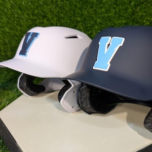May include: Two baseball helmets, one white and one navy blue, are displayed. Each helmet features a large letter "V" in a contrasting color. The white helmet has a dark blue "V", while the navy helmet has a light blue "V".