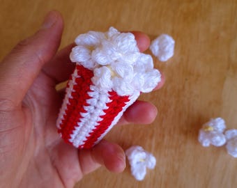 May include: A handmade crochet popcorn bucket with red and white stripes, filled with fluffy white popcorn. Several individual popcorn pieces are scattered around the bucket, all crafted with white yarn.