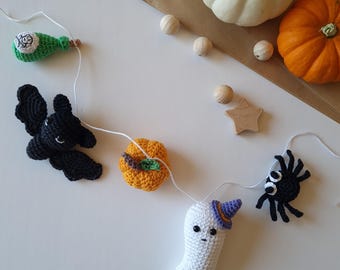May include: A handmade Halloween garland featuring crocheted figures: a green potion bottle, a black bat, an orange pumpkin, a white ghost with a purple hat, and a black spider. The garland is strung on white thread, with pumpkins and gourds in the background.