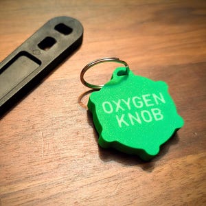 May include: A green, irregularly shaped keychain tag with the words "OXYGEN KNOB" in white lettering. A black tool is in the background. The items are on a wooden surface.