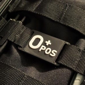 Puede incluir: Etiqueta rectangular negra con el texto blanco "O+ POS", sujeta a una correa negra. La etiqueta está colocada sobre una superficie negra texturizada, posiblemente una mochila. Se ve un cuchillo plateado.