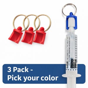 Op de afbeelding: Drie rode sleutelhangers met gouden ringen en een sleutelhanger met een blauwe dop. De spuit bevat een heldere vloeistof en bedrukte tekst. De blauwe banner toont de tekst "3 Pack - Pick your color."