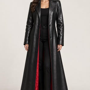 May include: A floor-length black coat with a pointed collar, long sleeves, and a front button closure. The coat features a vibrant red lining visible along the open front edges, contrasting with the smooth black exterior finish.