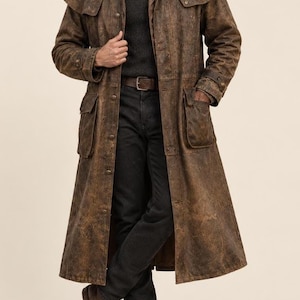 May include: A long, brown, distressed leather duster coat featuring a wide collar, a shoulder cape, and two large front patch pockets. The coat is fastened with metal buttons and has adjustable straps on the cuffs, worn over a dark gray sweater and jeans.