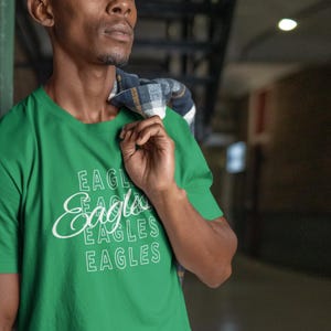 May include: A Kelly green t-shirt with the word "Eagles" in a white script and block font. The shirt is being worn by a person with a plaid shirt draped over their shoulder. The shirt is a solid color with the word "Eagles" repeated.