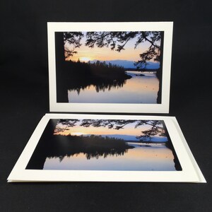 May include: Two white photo cards with a sunset over a lake with trees reflected in the water. The top card shows a close-up of the trees and the bottom card shows a wider view of the lake.