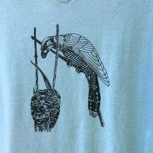 May include: A light blue t-shirt featuring a black and white illustration of a bird perched on a branch with a nest below.