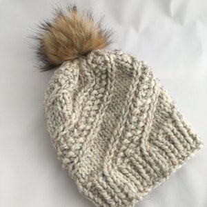 May include: A white knitted beanie with a brown faux fur pom pom. The beanie has a cable knit pattern.