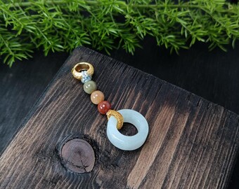 May include: A jade pendant with a light blue ring and a gold-colored accent. The pendant features a series of colorful beads in shades of green, orange, and red. The pendant is displayed on a wooden surface.