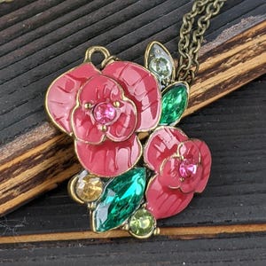 May include: A floral pendant necklace. The pendant features red enamel flowers with pink gemstone centres and green and yellow gemstone accents. The necklace hangs from a bronze-coloured chain, creating a vintage look.