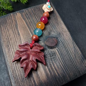 May include: A handcrafted wooden pendant featuring a carved maple leaf design, strung with a red cord and colorful beads. The pendant is displayed on a dark wooden surface, showcasing its intricate details and natural tones.