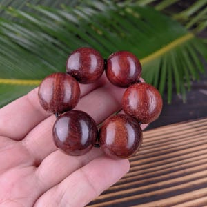 May include: A bracelet made of seven large, round, polished wooden beads. The beads are a deep reddish-brown color with visible wood grain. The bracelet is strung on a black cord. The background includes green leaves and a wooden surface.