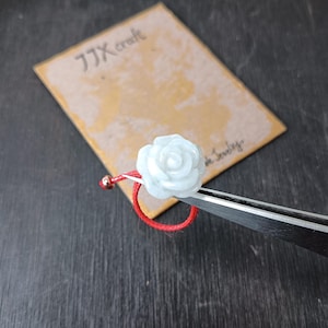 May include: A delicate ring featuring a white rose-shaped gemstone set on a red cord band. The ring is held by tweezers, with a small gold bead accent. A tan card with the text "JJX craft" is in the background.