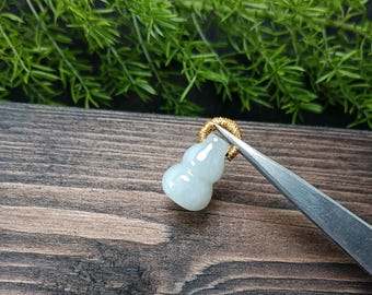 May include: A pale green jade gourd pendant with a gold-colored loop. The gourd is smooth and polished, with a slight translucence. The pendant is held by a pair of tweezers against a wooden surface with green foliage in the background.