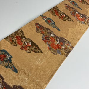 Può includere: Tessuto decorativo con sfondo dorato e motivi ripetuti di uccelli stilizzati e disegni floreali in rosso, marrone e blu. Il tessuto ha un aspetto strutturato e viene probabilmente utilizzato per l'arredamento della casa o l'artigianato.