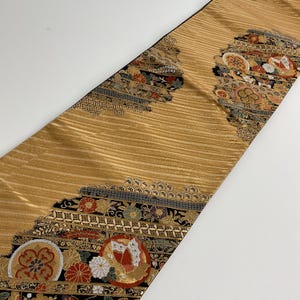 Può includere: Un tessuto color oro con un motivo strutturato e a righe. I bordi sono decorati con disegni geometrici neri e grigi e motivi floreali e circolari colorati. Il tessuto è probabilmente un oggetto decorativo, come un runner da tavolo o una fascia decorativa.