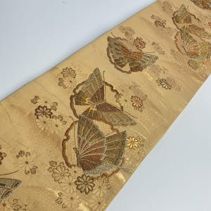 Può includere: Un tessuto decorativo con un motivo ripetuto di farfalle e fiori. Le farfalle hanno motivi alari dettagliati in tonalità di marrone e oro, su uno sfondo oro chiaro. I fiori sono anch'essi in oro e marrone.