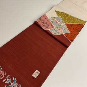 以下が含まれることがあります： 深い赤色の背景を持つ、長方形の織物。上端には、ピンク、グレー、オレンジの幾何学模様のパッチワークデザインがあります。下部には、小さな長方形の日本の文字のラベルが見えます。この織物は装飾品である可能性があります。