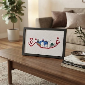 May include: A framed cross-stitch embroidery featuring a stylized red Arabic script outline filled with a colorful cityscape illustration. The design includes white buildings, a green domed structure, and blue elements, all set against a white fabric background.