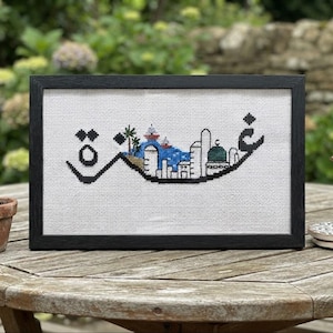 May include: A framed cross-stitch embroidery on white fabric featuring black Arabic calligraphy that forms a stylized cityscape. The design includes blue water, green palm trees, and a green domed building integrated into the black lettering.