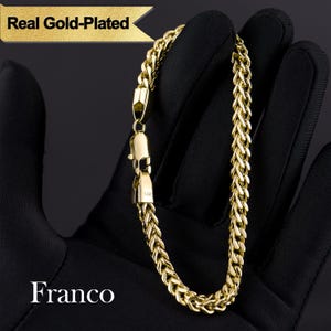 May include: A gold-plated Franco bracelet is displayed against a black background. The bracelet features a woven chain design with a secure clasp. The text "Real Gold-Plated" and "Franco" are visible.