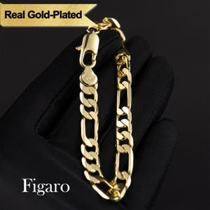 May include: A gold-plated Figaro chain bracelet. The bracelet features a classic curb link design with alternating long and short links. The clasp is a lobster claw style. The text "Real Gold-Plated" and "Figaro" are visible.