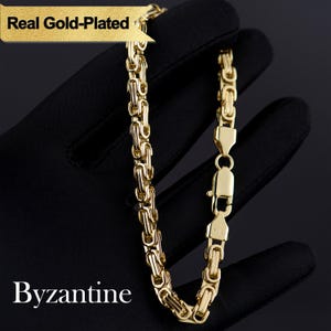 May include: A gold-plated Byzantine chain bracelet. The bracelet is made of interlocking links and features a secure clasp. The text "Real Gold-Plated" and "Byzantine" are visible in the image. The bracelet is displayed against a black background.
