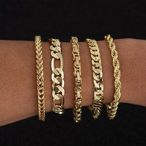 May include: Five gold-colored bracelets with different chain designs are displayed on a wrist against a black background. The bracelets include a herringbone, Figaro, Byzantine, curb, and rope chain style.