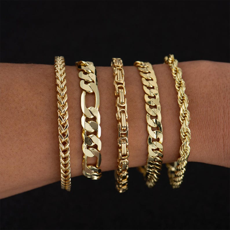 May include: Five gold-colored bracelets with different chain designs are displayed on a wrist against a black background. The bracelets include a herringbone, Figaro, Byzantine, curb, and rope chain style.