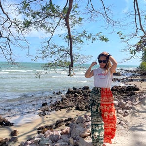 May include: A woman wearing a white tank top with the text "imissyou" in Thai script, and green and orange patterned pants with elephants and palm trees. She is standing on a sandy beach with rocks and the ocean in the background.