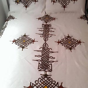 May include: White bed linens with intricate brown and yellow embroidered patterns. The design features geometric shapes and knot-like details, creating a decorative and textured appearance. The set includes pillowcases and a bedspread.